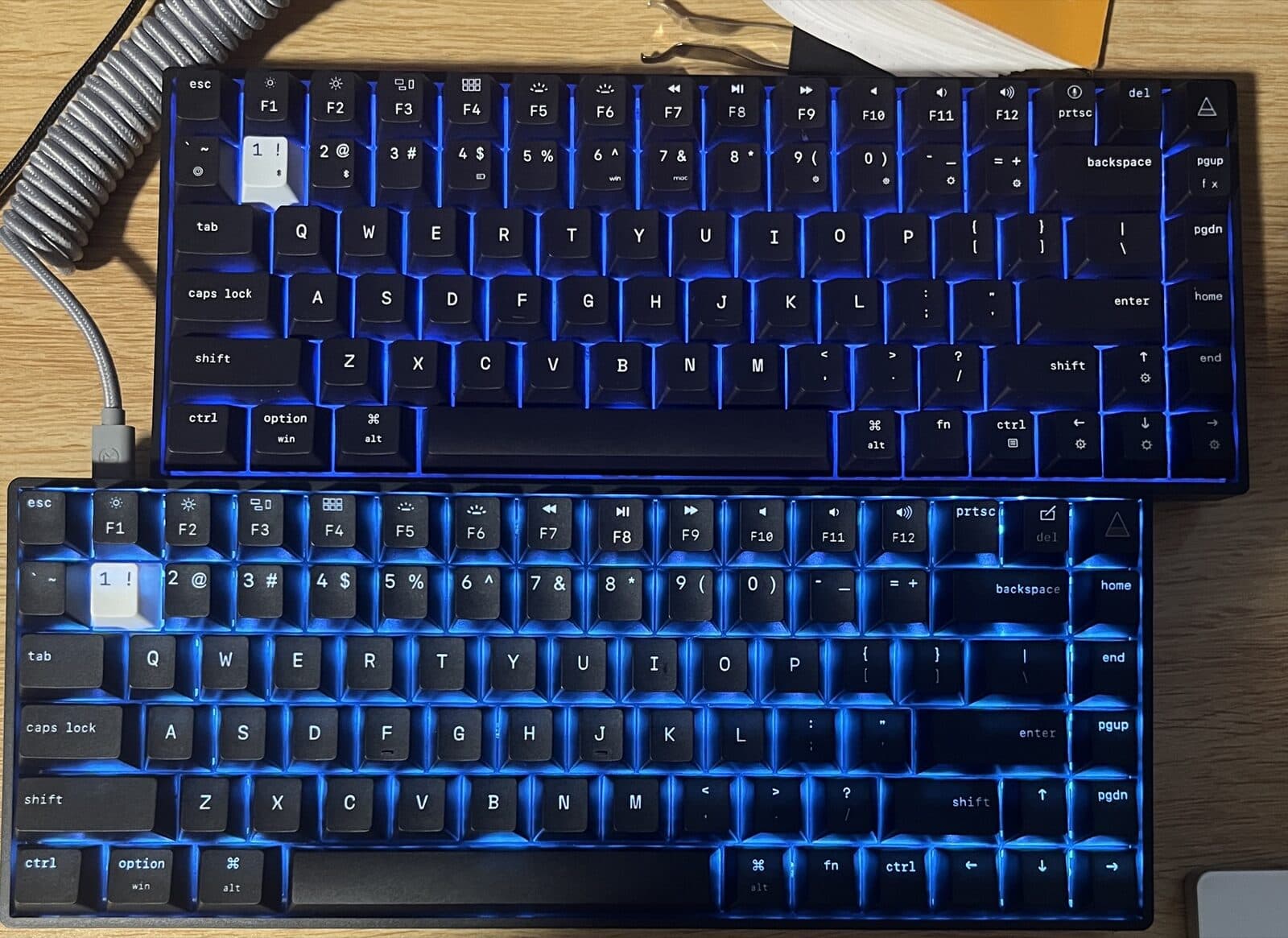The same two keyboards in daylight on a wooden desk, with a coiled grey cable. Black keycaps with white legends, blue backlight showing between the keys.