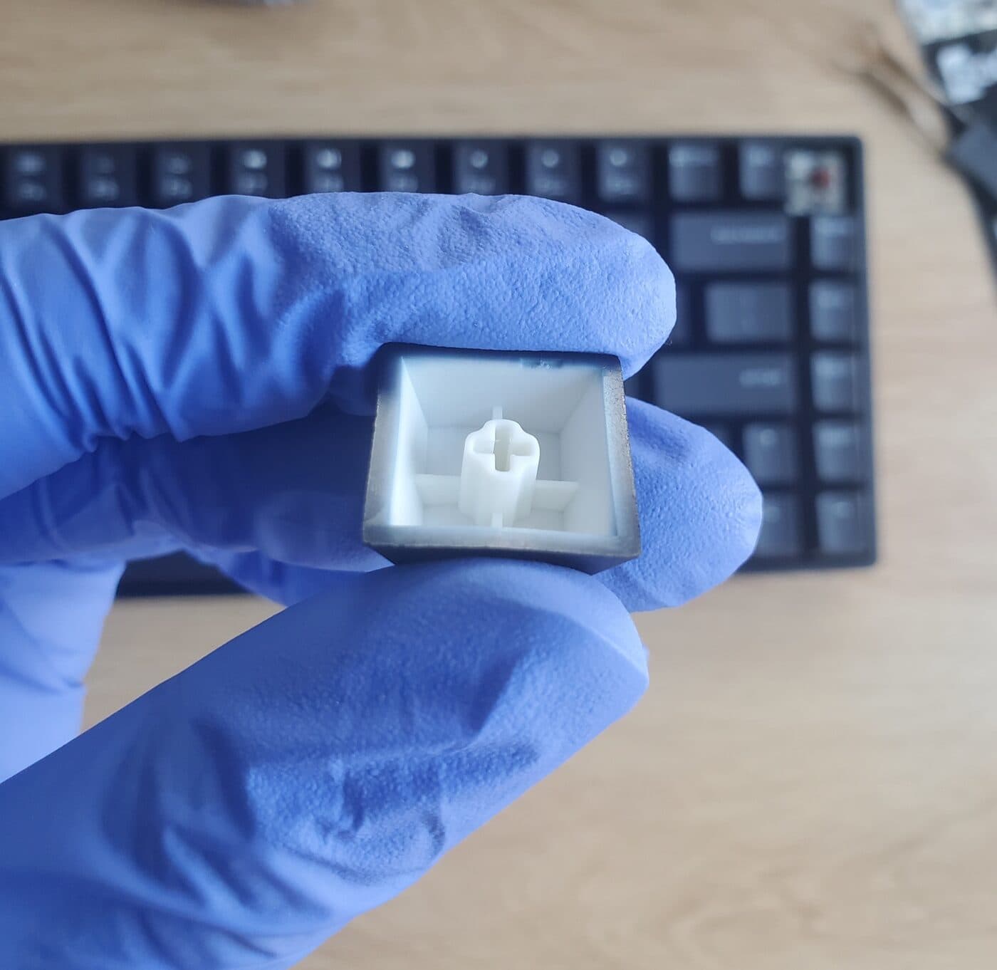 A single black keycap held upside down in a gloved hand, showing the thick white PBT walls and the stem.
