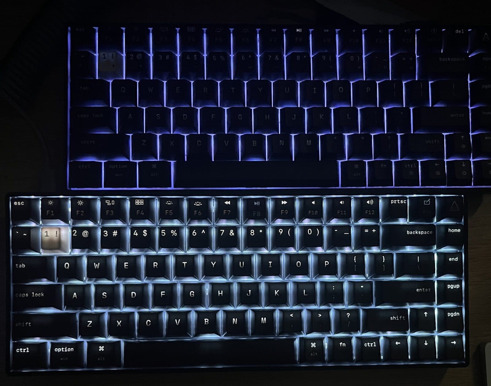Two Code One keyboards in the dark. The earlier board on top shows light leaking around the keycaps but the legends barely read. The final board below has every legend lit clearly.